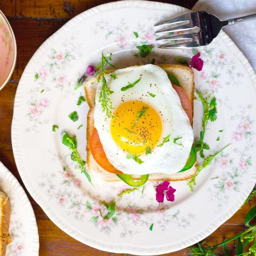 Een bord met twee sneetjes koolhydraatarm brood belegd met ham, kaas, tomaat, avocado en sla, afgewerkt met een gebakken spiegelei en een vleugje paprikapoeder.