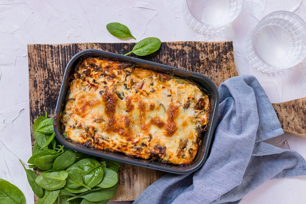 Ovenschaal met goudbruin gegratineerde lasagne op een houten plank, omringd door verse spinazieblaadjes en glazen water, met een blauwe linnen servet.