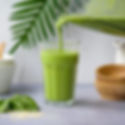 Glas groene smoothie van spinazie, banaan en avocado, vers gegoten uit een blender, met spinaziebladeren en sesamzaadjes op tafel.