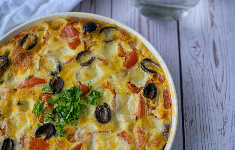 Ovenschaal met goudbruine quiche gevuld met champignons, olijven en tomaat, afgewerkt met peterselie.