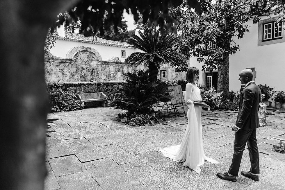 A couple stands facing each other in a courtyard with stone tiles and lush greenery. The woman wears a long dress, the man a suit. The mood is intimate.