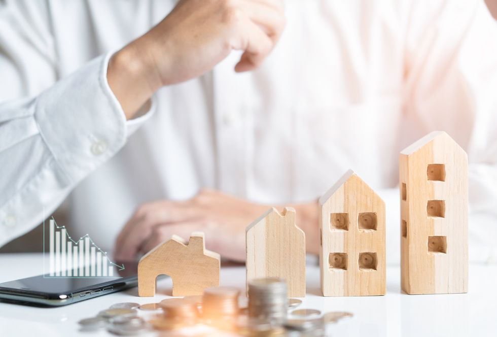 Person in weißem Hemd sitzt an einem Tisch mit Holzmodellen von Häusern, Münzstapeln und einem Smartphone, daneben ein eingeblendetes Balkendiagramm.
