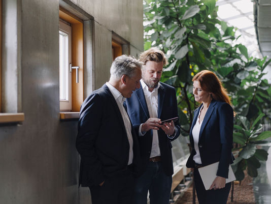 Drei Personen in Businesskleidung stehen in einem modernen Büroflur und betrachten gemeinsam ein Smartphone.
