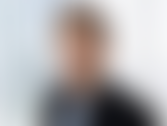 Porträtaufnahme von Autor Marek Fritz in einem schwarzen Sakko und hellblauem Hemd, vor einem hellen Hintergrund mit großen Fenstern und unscharfer Stadtansicht.