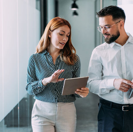 Zwei Personen gehen durch einen modernen Büroflur und betrachten gemeinsam ein Tablet.