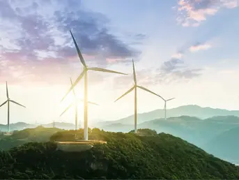 Mehrere Windkraftanlagen auf grünen Hügeln bei Sonnenaufgang, mit Bergen im Hintergrund und leicht bewölktem Himmel.