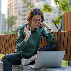 Smiling young woman waving hello and doing remote work video call at park bench_edited_edi