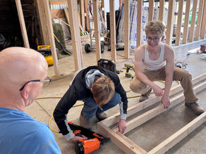 Three people building a wooden frame in a workshop. One uses an orange nail gun. Tools and wood are scattered around. Bright, sunny atmosphere.