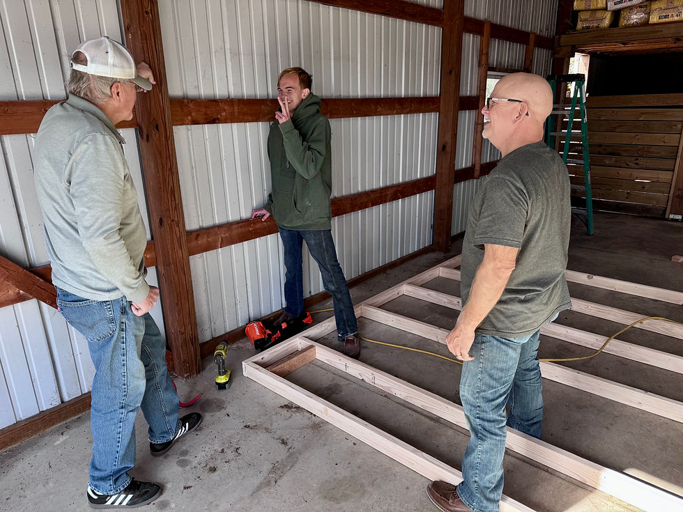 Three men in a garage discuss construction plans, with wooden beams on the floor. One leans on the wall, tools scattered around.