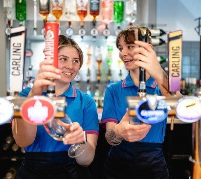 Two smiling Parkdean Resorts bar staff in blue uniform polo shirts pour pints from the beer taps behind a holiday park bar.