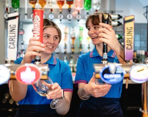 Two smiling Parkdean Resorts bar staff in blue uniform polo shirts pour pints from the beer taps behind a holiday park bar.