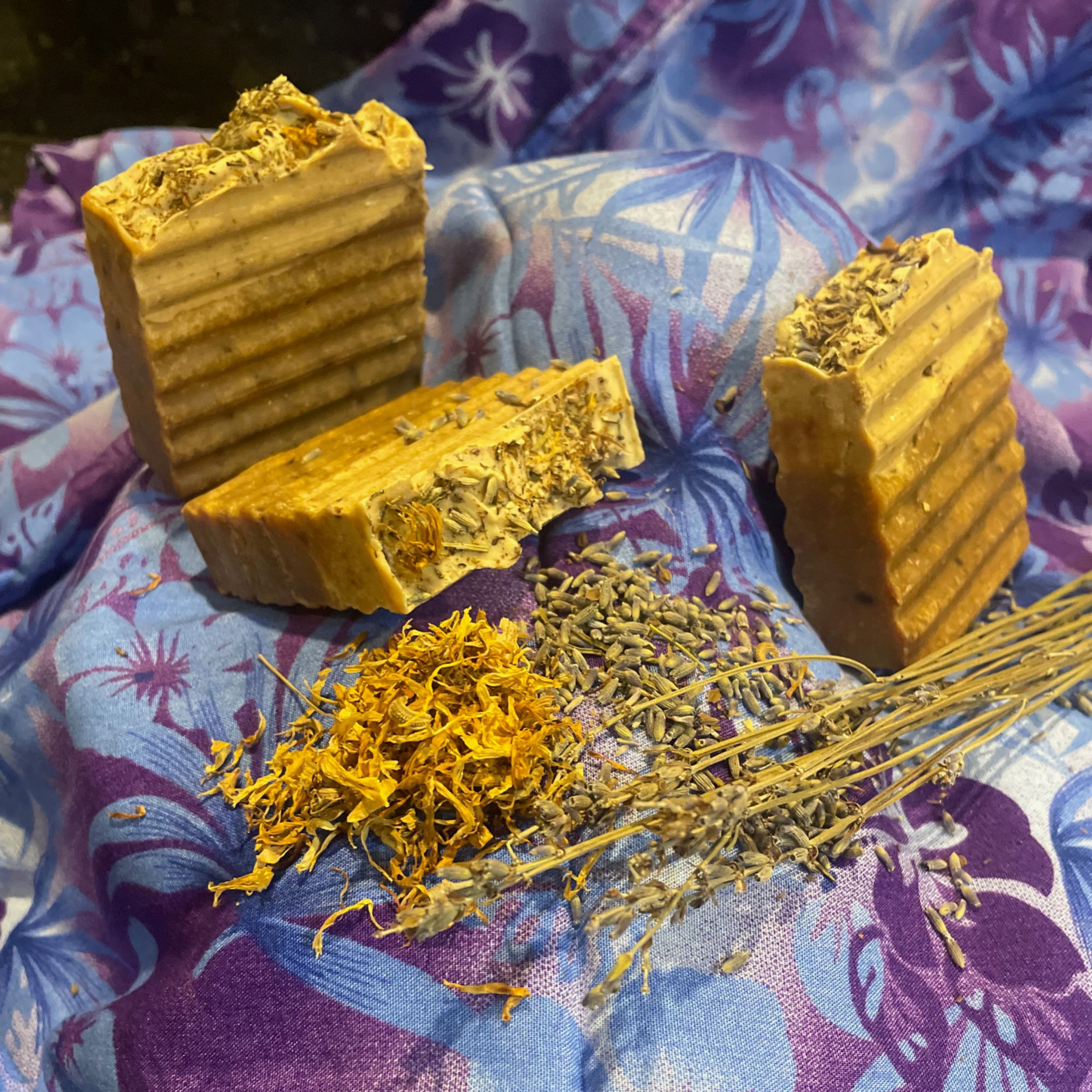 soaps laying on purple textured cloth surrounded by herbs and on them