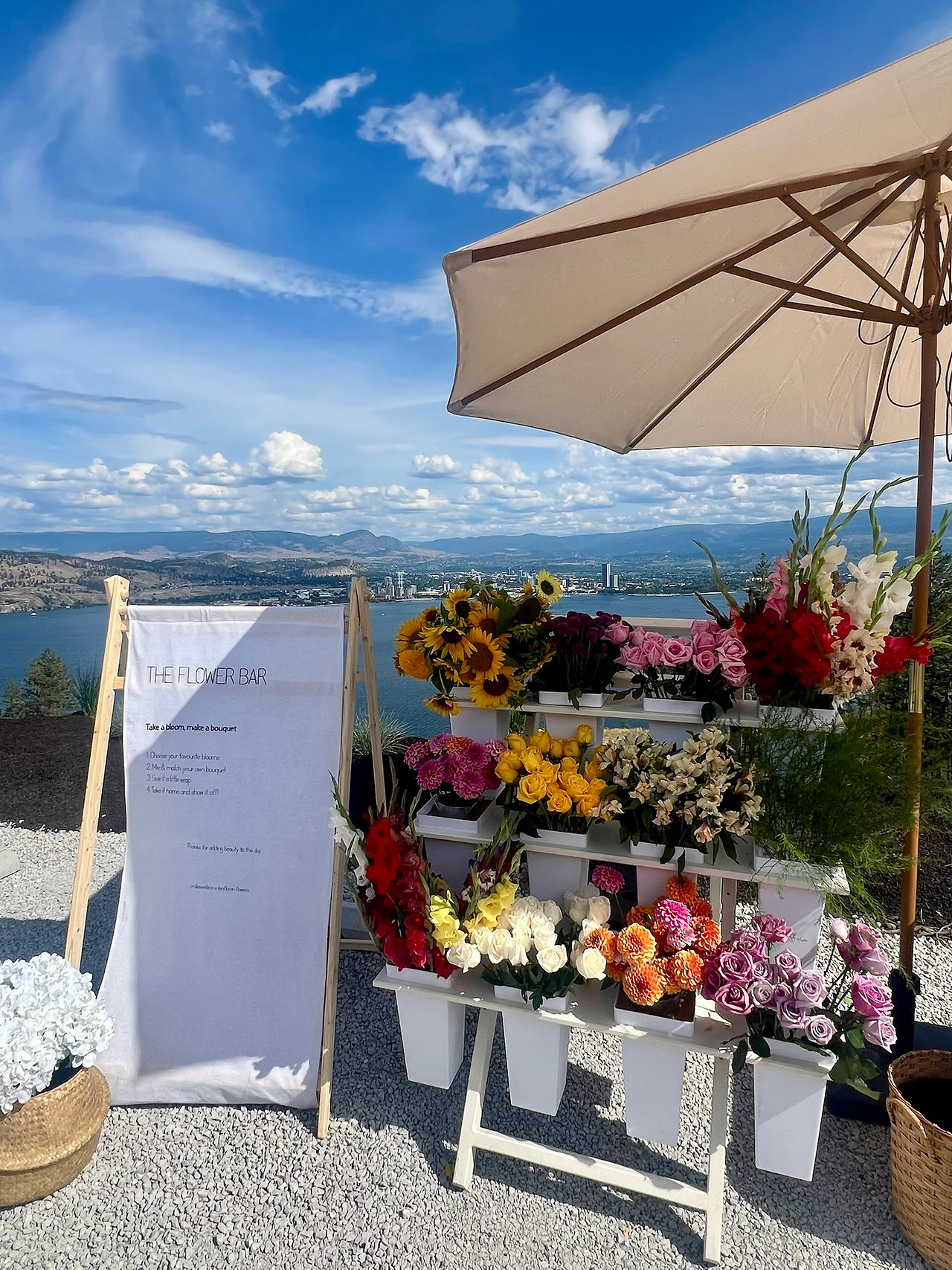 flower bar experience set up at event with Okanagan lake views. Flower bar displays variety of colourful flowers, a linen flower bar sign, and an umbrella.
