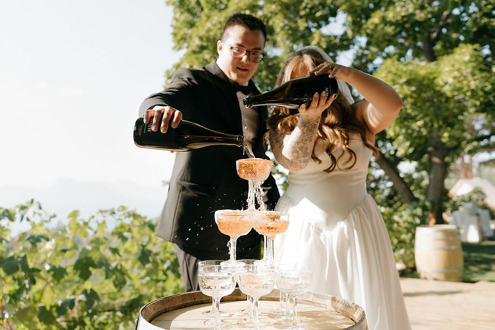 Bride and groom in wedding attire pour champagne into a champagne tower of glasses outdoors at Penticton wedding venue, N1 Estate Farm. Sunlit greenery and barrels in the background.