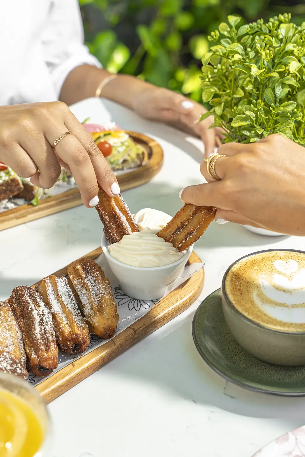 Two customers dipping artisanal stuffed churros into cream, with a cappuccino and vibrant green plants in the background