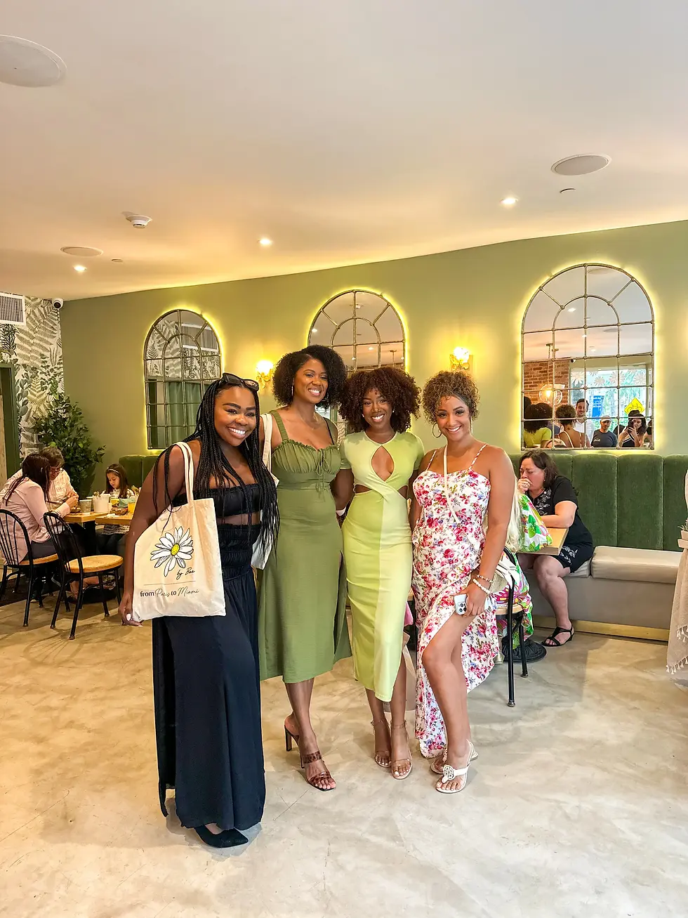 Four women posing in colorful dresses inside a green café with mirrors and soft lighting