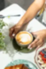 Hands holding a latte with leaf foam art beside a croissant and vibrant fruit bowl on a sunlit cafe table at Café Bastille