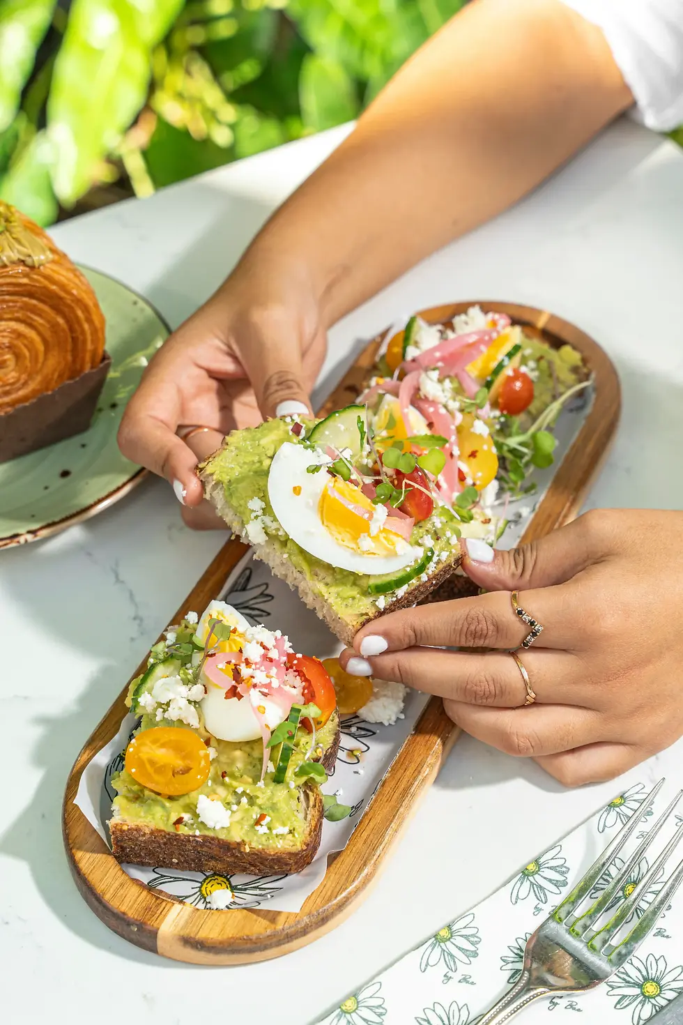 Hands holding avocado toast topped with boiled egg, cherry tomatoes, feta, and microgreens on an outdoor cafe table
