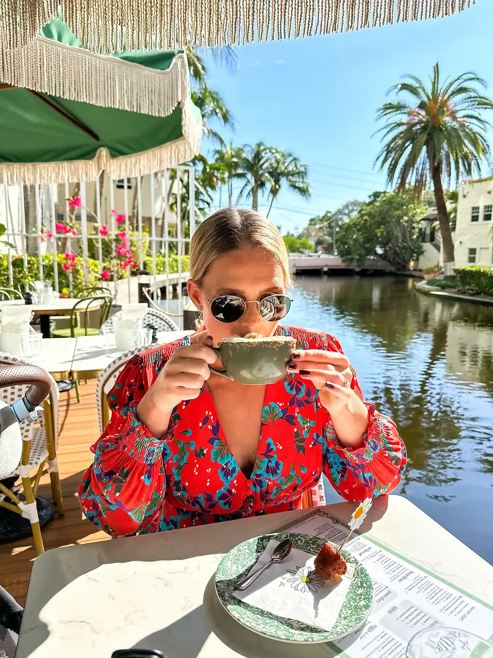 Woman in a floral dress sipping a cappuccino at a riverside patio with green fringed umbrellas and surrounded by palm trees