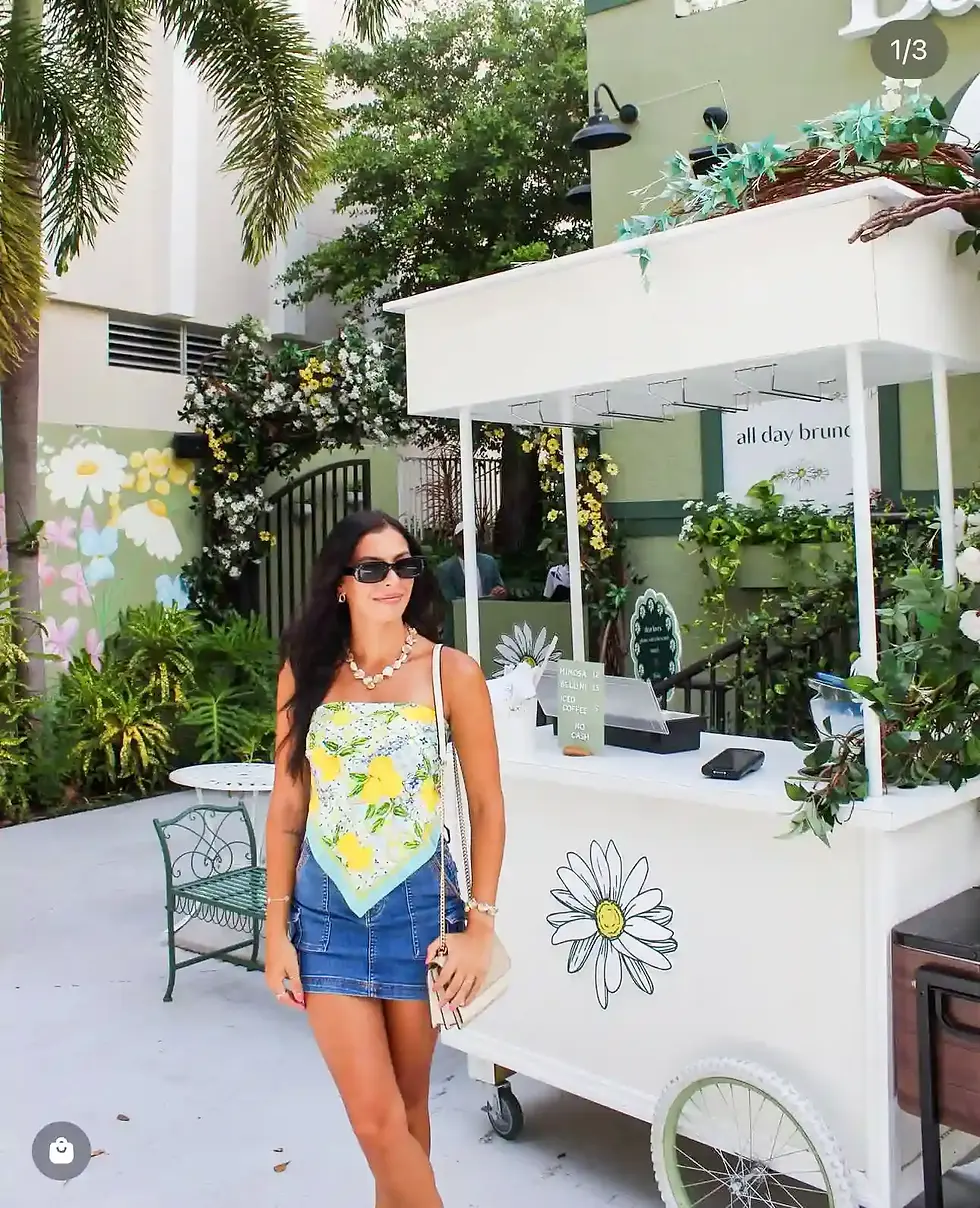 Smiling woman wearing denim skirt and floral top near a white flower-decorated cart at the café entrance