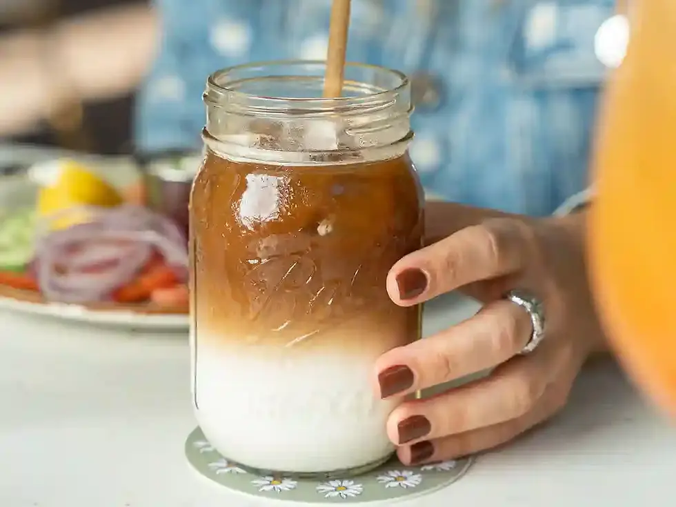 An iced latte in a tall glass jar with the espresso settling down through the milk, held over a brunch table.