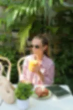 A customer in floral blouse sipping an iced drink at a cafe table with a croissant, notebook, and potted plants.