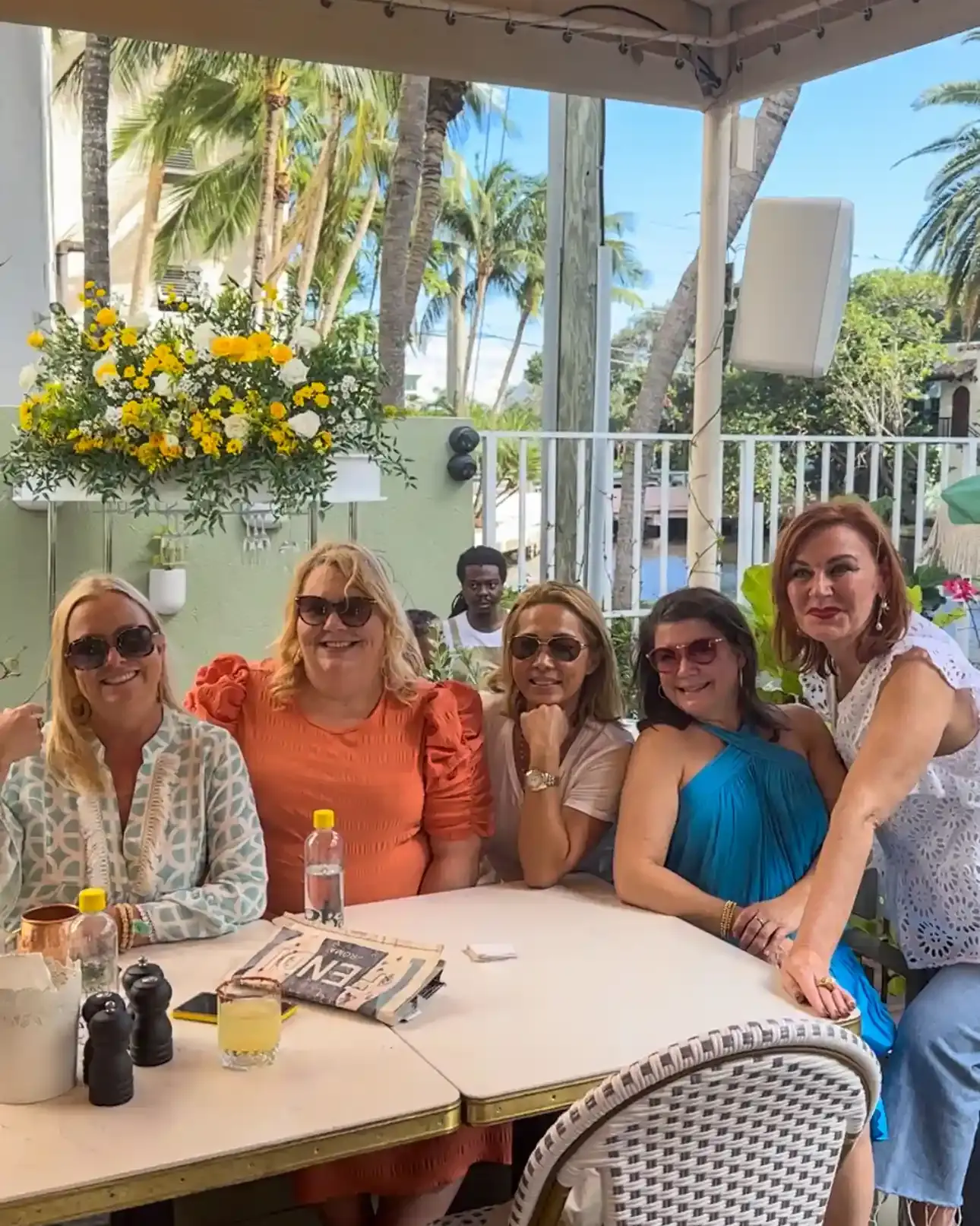Five guests seated together at an outdoor table at Cafe Bastille Fort Lauderdale, with a yellow and white flower arrangement, palm trees and the canal behind them.