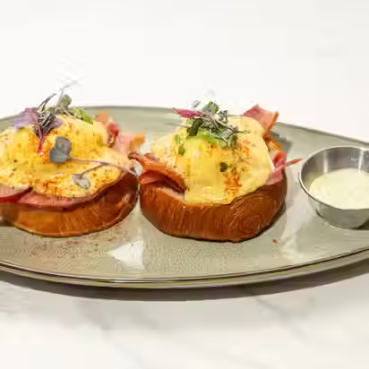 Two open-faced Country Ham Benedicts on toasted brioche topped with paprika-dusted hollandaise, microgreens, and salt-cured country ham, served on a gray ceramic plate with a small ramekin of cream sauce
