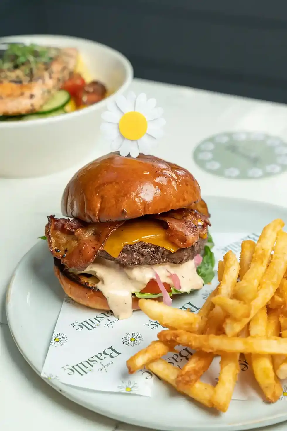 Close-up of a bacon cheeseburger with secret sauce and fries on a plate, topped with a small decorative daisy pick
