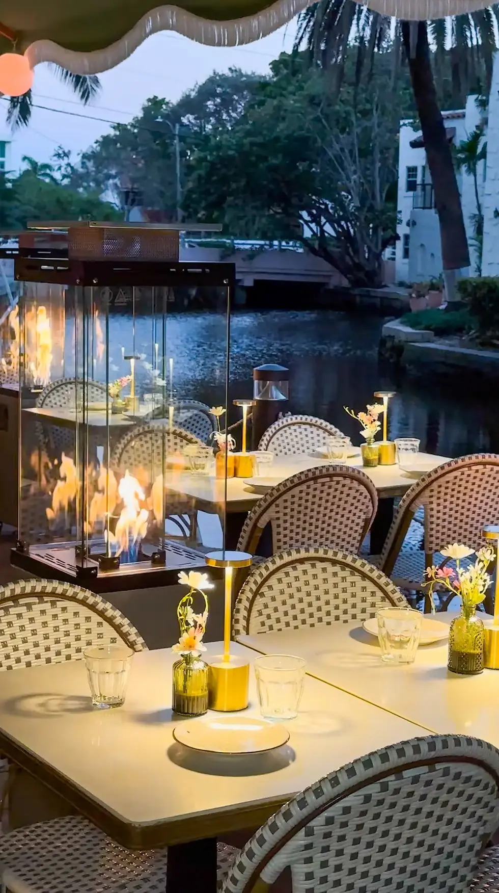 An outdoor dining area with lit fire lamps, plants, and tables by a canal, with a warm and inviting atmosphere.