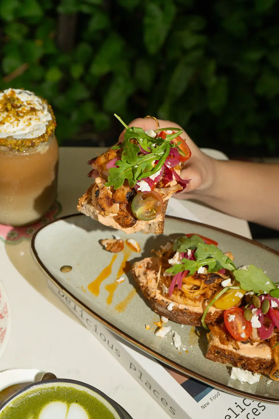 A slice of Harvest Chicken Toast being lifted from a shared plate with arugula, pickled onions, and tomatoes on the table