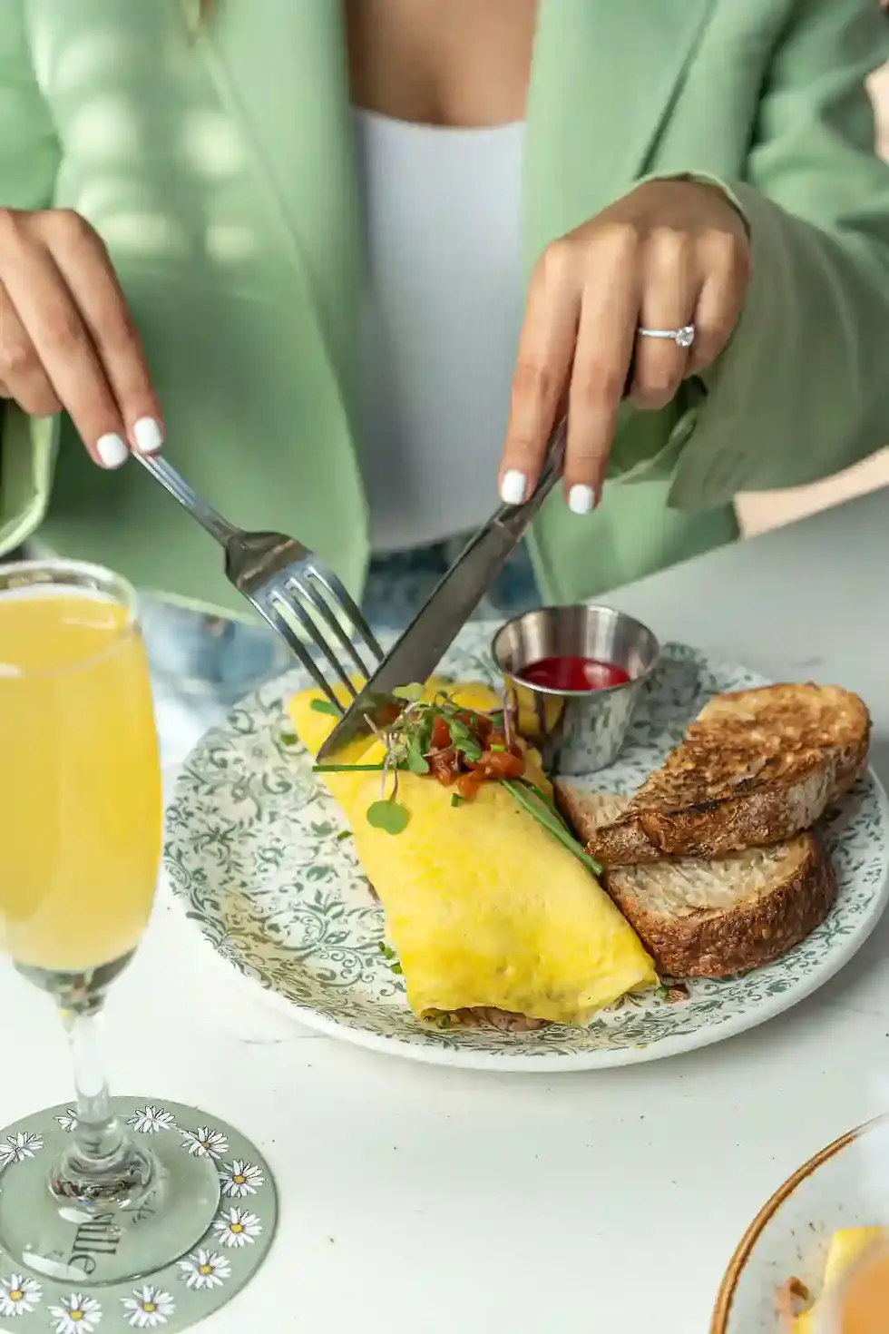 Woman in a green blazer, cutting into the Parisian Omelette Platter, served with toasted bread and a side of sauce, alongside a mimosa