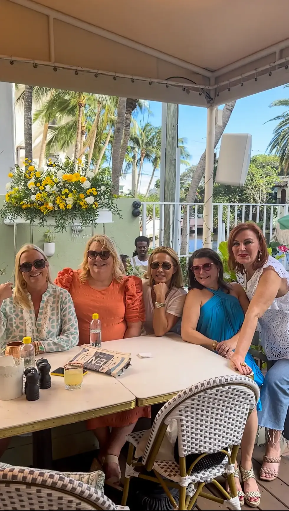  Group of friends at Café Bastille enjoying brunch under a canopy with tropical plants and flowers, most wearing sunglasses.