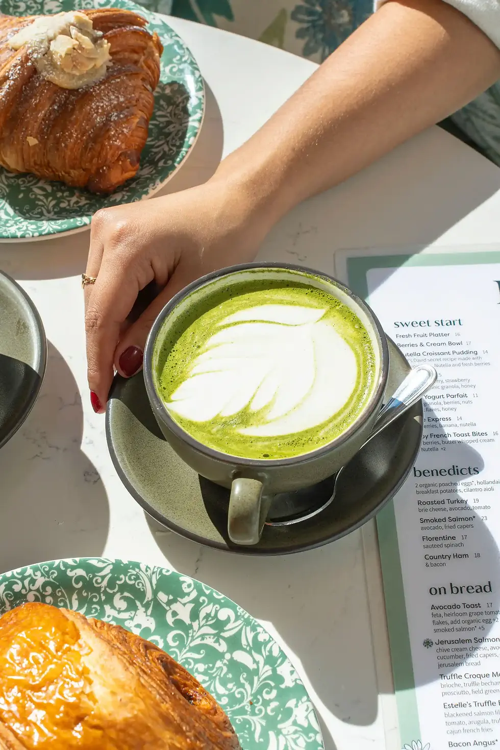 A hand holds a matcha latte beside croissants and a brunch menu on a white table at a Las Olas brunch cafe