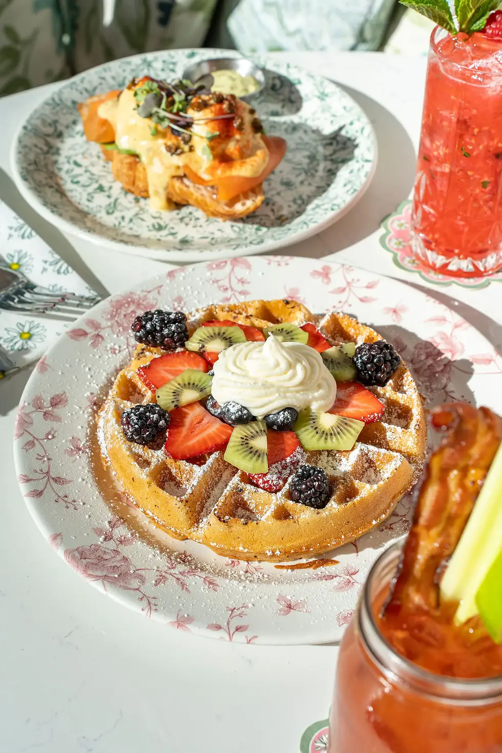 Golden waffle topped with whipped cream, sliced strawberries, kiwi, blackberries, and powdered sugar on floral china.