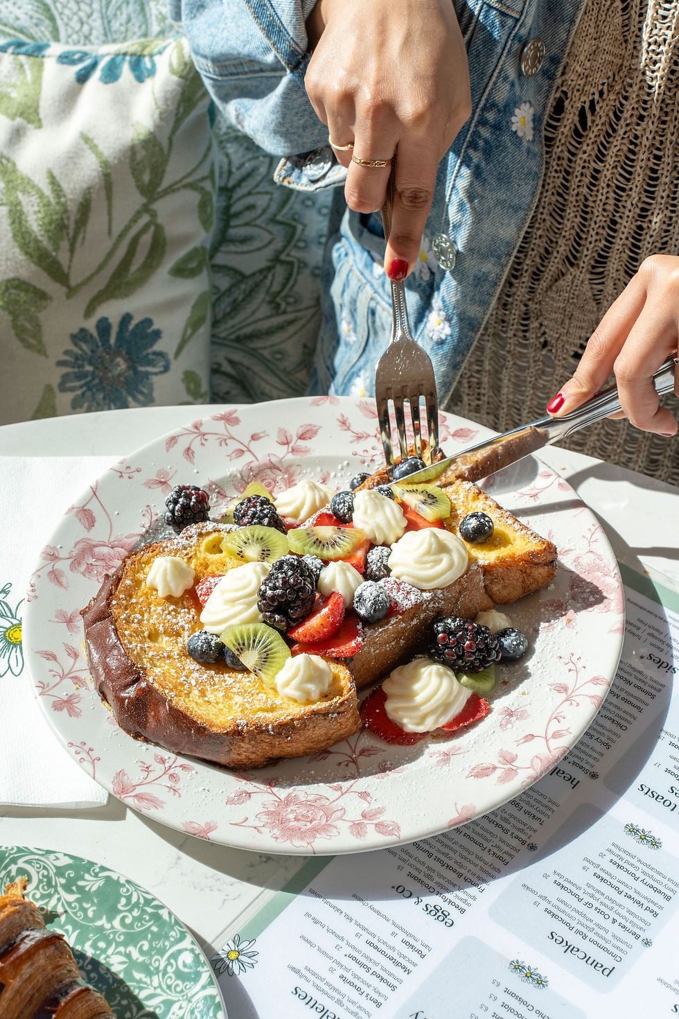 A guest cutting into The Classic French Toast topped with kiwi, strawberries, blackberries, blueberries and piped butter whipped cream on pink floral china.