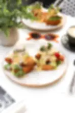 Two chicken benedict servings topped with greens and hollandaise on a plate with sunglasses, coffee, and a plant on the table