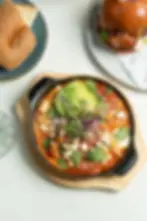 Bae's Shakshuka served with organic eggs, feta, a Jerusalem bagel, and sliced avocado, served on a skillet and wooden board.