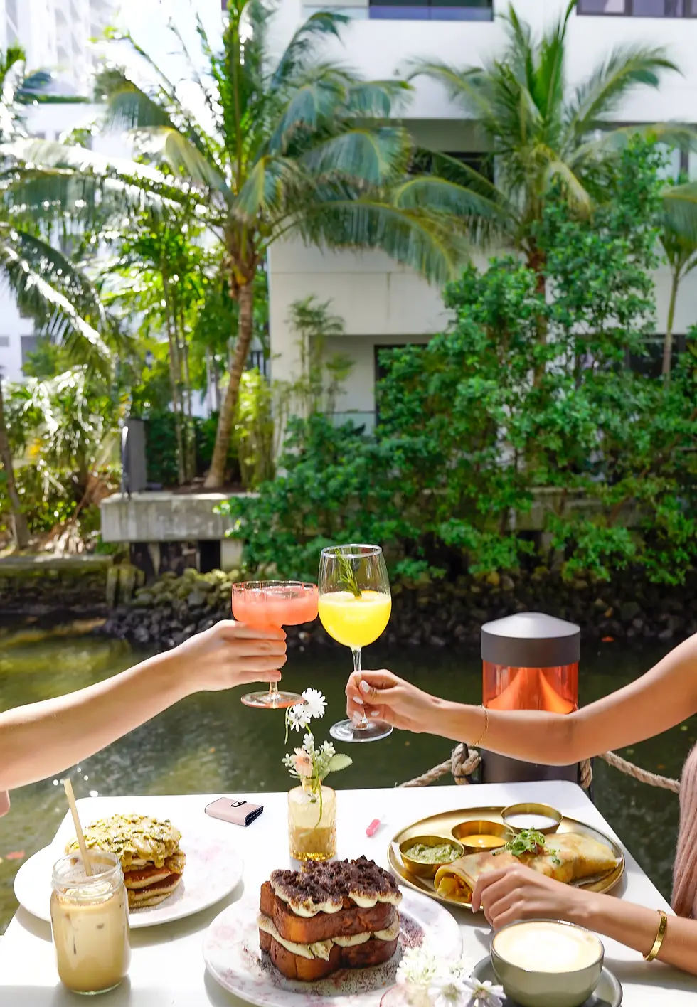 A brunch table with pancakes, toast, cocktails, and coffee by the water surrounded by greenery at Cafe Bastille.