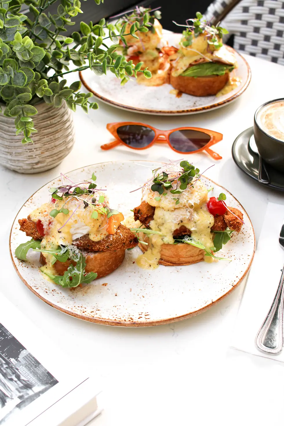 Chicken Milanese Benedict with poached eggs, microgreens, hollandaise, arugula, and tomato, served at a bright cafe table