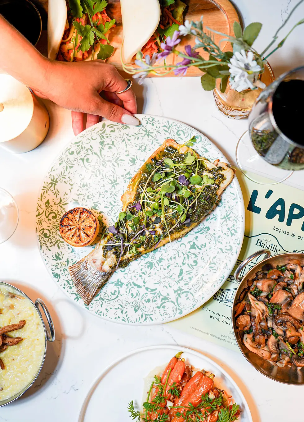 A plate of branzino topped with microgreens, alongside roasted lemon and other dishes in the background.