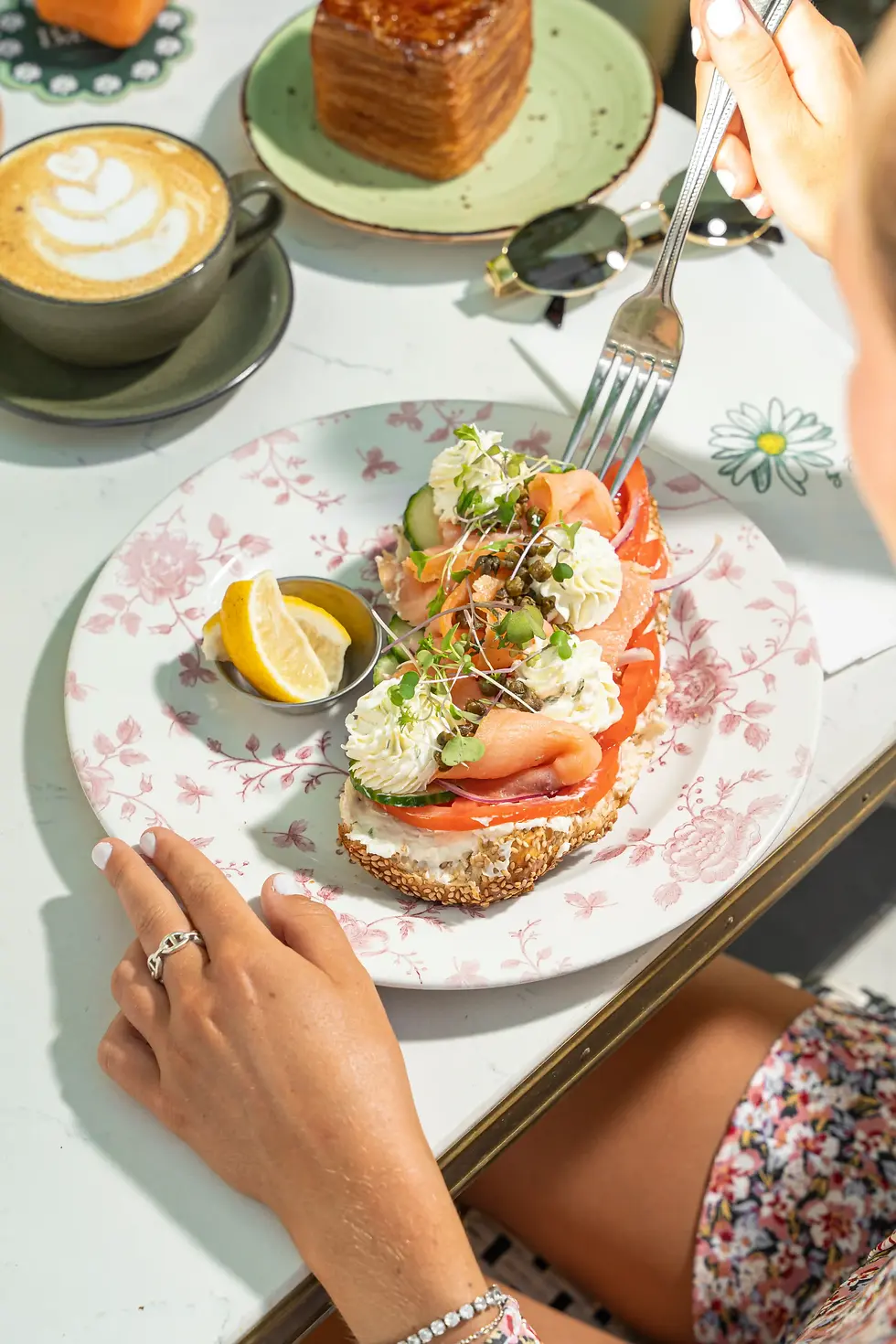 Person enjoying Mediterranean-style salmon lox toast with chive cream cheese, tomato, cucumber, and capers at an outdoor cafe