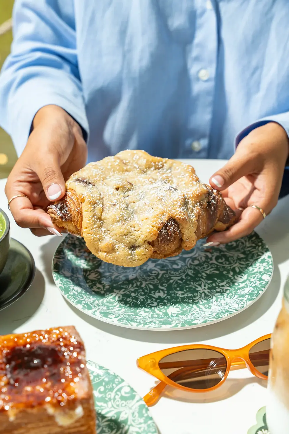 hands holding a sugar-dusted crookie croissant with chocolate chip cookie layers over a green plate
