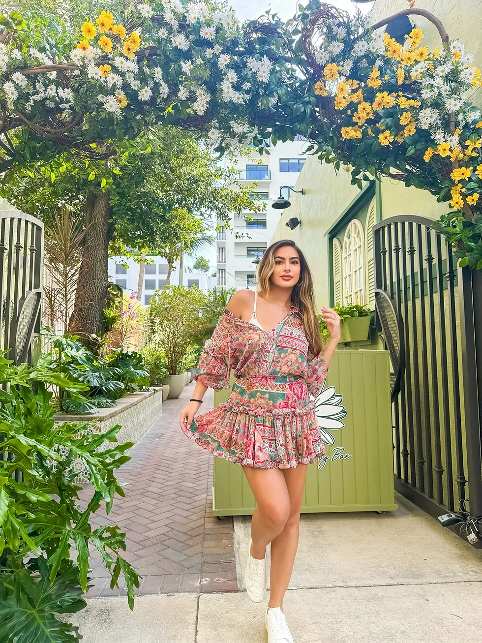 Woman in a floral boho dress posing under an archway of yellow and white flowers at the outdoor cafe entrance.