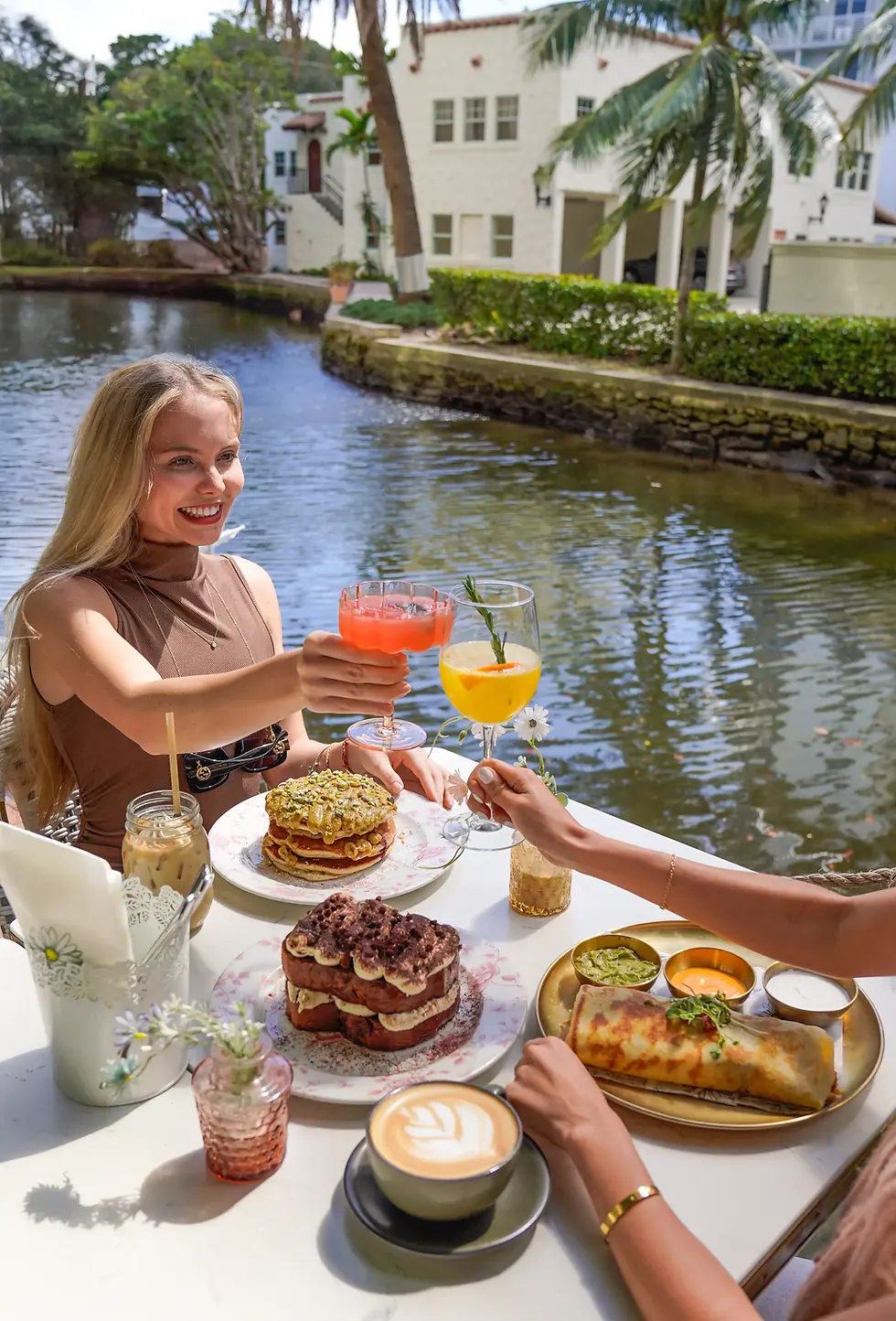 Women toasting with colorful cocktails over a brunch table by the water, with pancakes, tiramisu toast, crepe, and coffee.