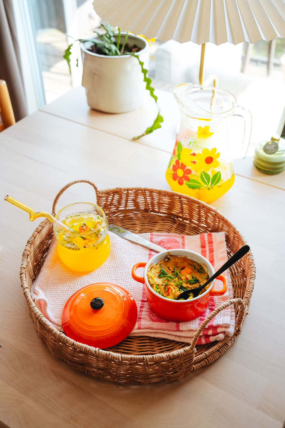 Wicker serving tray on a table with a mini vegan tofu frittata in an orange ramekin and refreshing golden watermelon drink. Plant and lamp in the background, creating a cozy dining atmosphere.