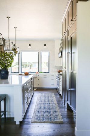 A bright and airy kitchen featuring a white island with a blue vase of greenery and upholstered bar stools. The space is complemented by dark wood cabinetry on one side, offering a rich contrast. Large windows at the end of the kitchen provide natural light, while pendant lights with metal frames add a modern touch. A soft, patterned runner rug stretches along the dark wood flooring, adding warmth and texture to the clean, functional design. Designed by Bar Kay Design Co., an interior design business located in Oregon, specializing in services across Eugene, Bend, and Portland, showcasing high-quality craftsmanship and innovative design.