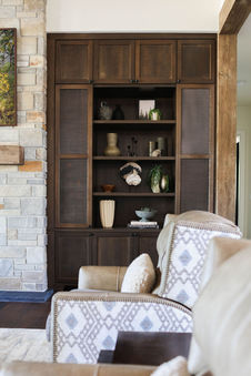 A close-up view of a dark wood built-in shelving unit, featuring a mix of decorative vases, books, and greenery arranged elegantly. The shelving is flanked by a stone fireplace on one side and framed by rustic wood beams, adding texture and warmth to the space. In the foreground, patterned armchairs with neutral tones complement the cohesive and inviting design. Designed by Bar Kay Design Co., an interior design business located in Oregon, specializing in services across Eugene, Bend, and Portland, showcasing high-quality craftsmanship and innovative design.