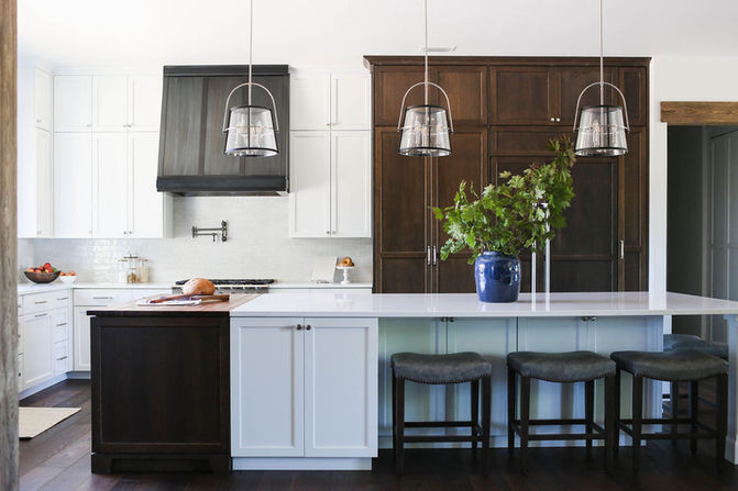 A modern kitchen featuring a large white island with a blue vase filled with greenery, paired with upholstered bar stools. The kitchen includes a mix of white cabinetry and dark wood cabinets, creating a striking contrast. A sleek black range hood is mounted above the stove with a pot filler faucet, adding both functionality and style. Two pendant lights with metal frames hang above the island, providing soft lighting. The overall design blends modern elegance with warm, natural tones. Designed by Bar Kay Design Co., an interior design business located in Oregon, specializing in services across Eugene, Bend, and Portland, showcasing high-quality craftsmanship and innovative design.