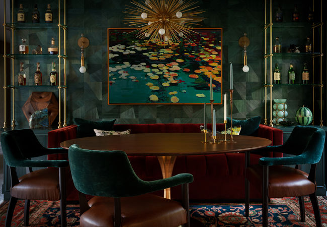 A luxurious dining room featuring a round wooden table, deep green velvet chairs, and a rich red banquette. The space is adorned with a striking lily pad artwork, a dramatic starburst chandelier, and brass sconces, creating a moody yet elegant atmosphere. Designed by Bar Kay Design Co., an interior design business located in Oregon, specializing in services across Eugene, Bend, and Portland, showcasing high-quality craftsmanship and innovative design.